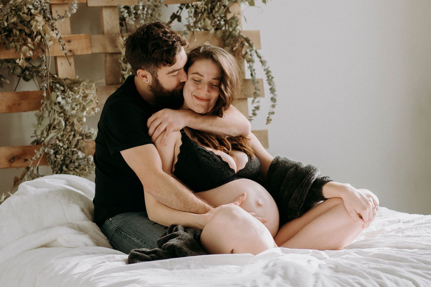 Tender moment between couple on white bedding with wooden accent wall.