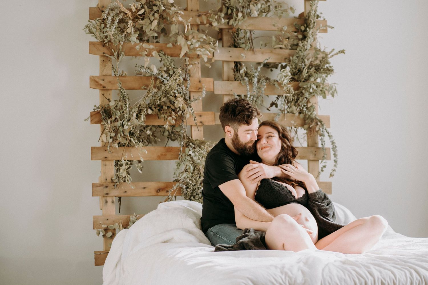 Couple cuddling on white bed against wooden lattice wall with dried botanical decor.