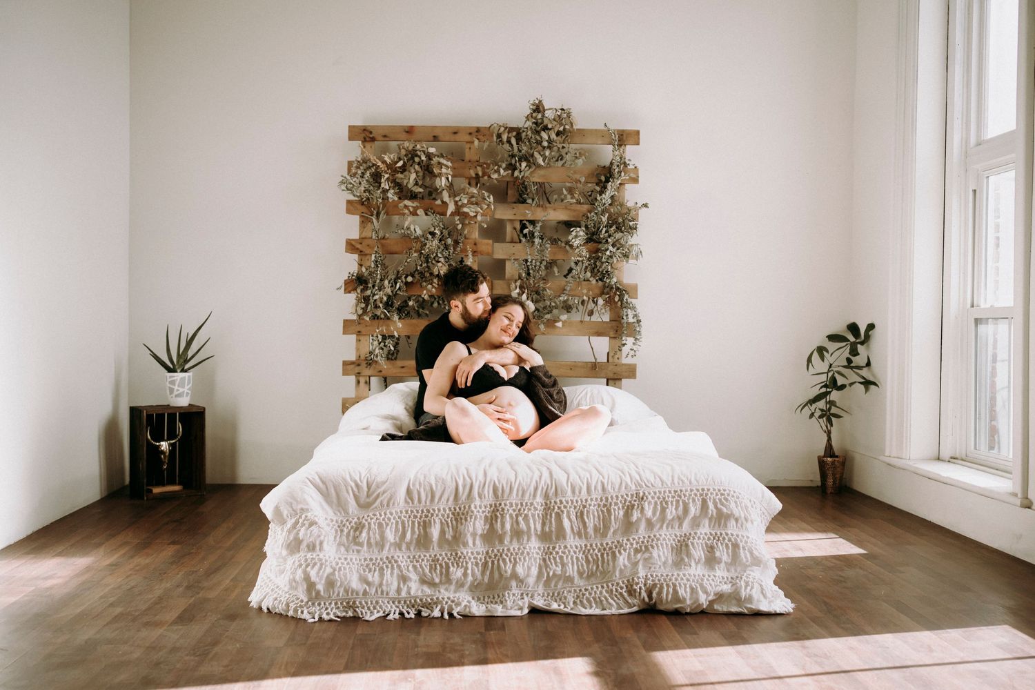 Intimate portrait on white bed with wooden wall accent and plant decor.