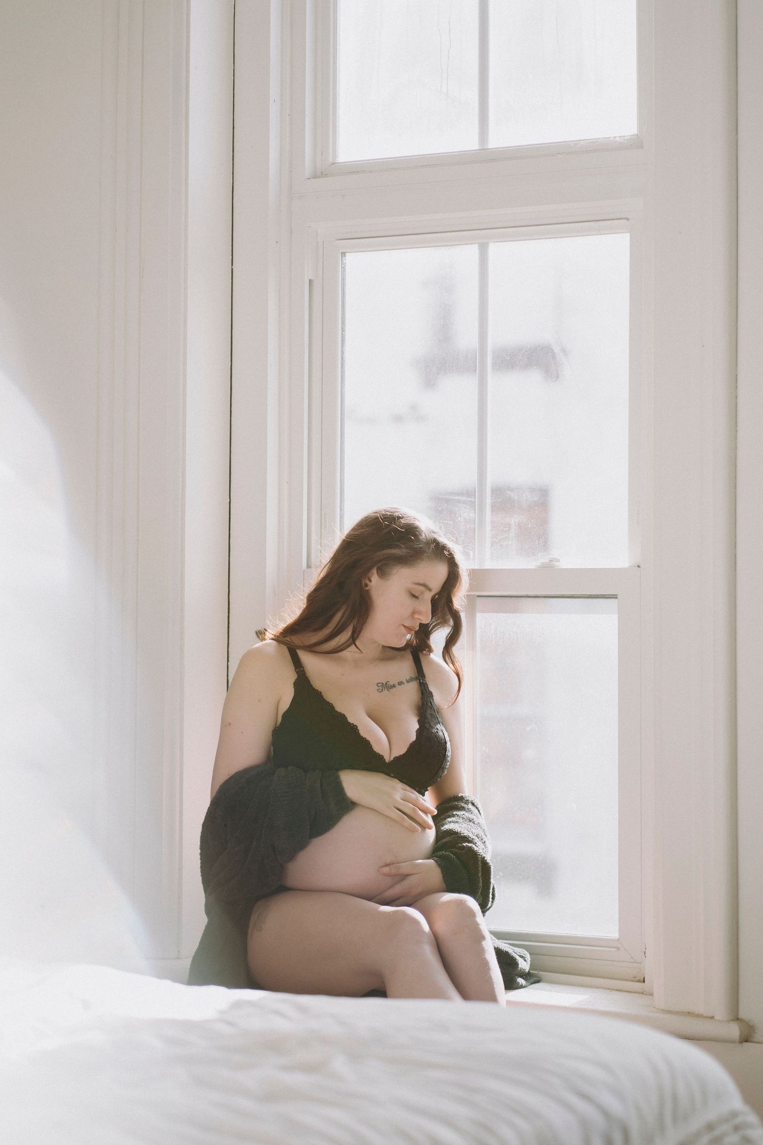 A soft maternity photoshoot by a bright window with a woman sitting in black lingerie.