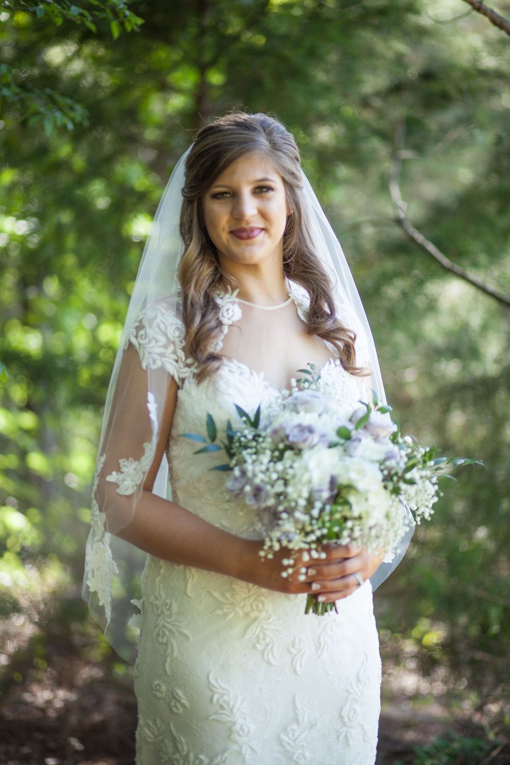 A bride in a detailed lace gown holds a delicate bouquet of white flowers in a wooded setting.