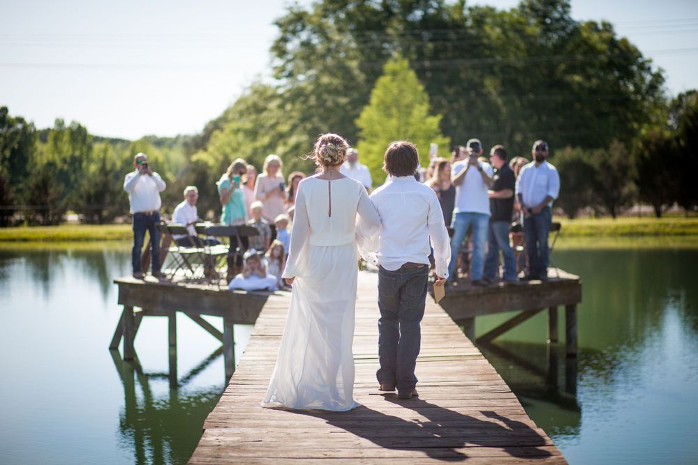 A couple walks along a wooden dock over a lake surrounded by wedding guests on a sunny day.
