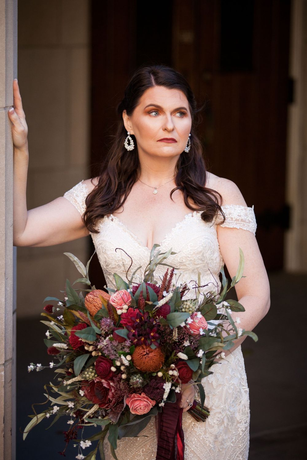 An elegant bride in a lace wedding dress holds a burgundy and blush bouquet against a stone column.