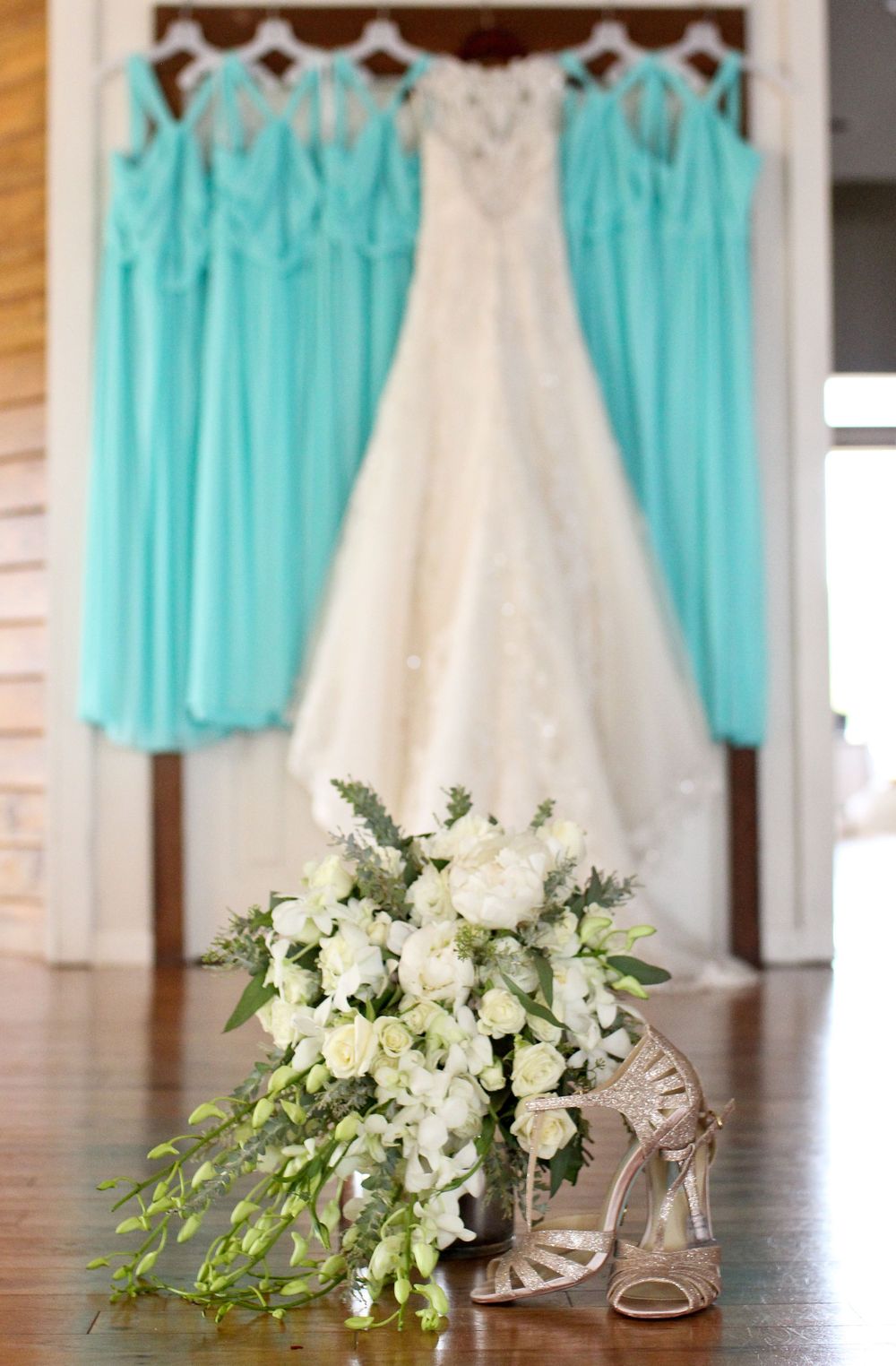 White bridal gown hangs with turquoise bridesmaid dresses above a cascading white floral bouquet and shoes.
