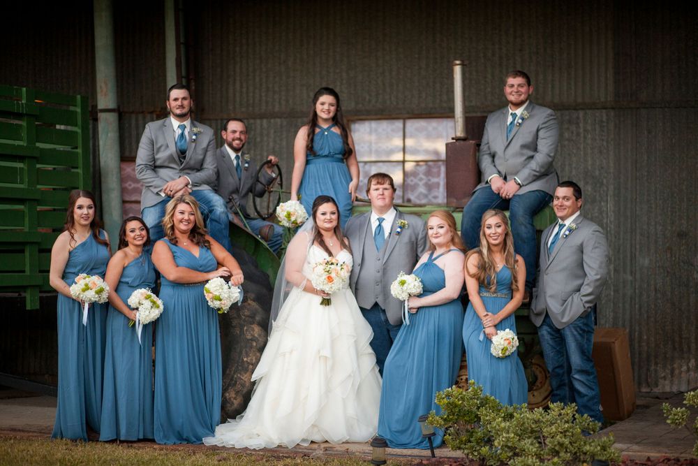 Wedding party in blue and gray attire poses together outside rustic barn.