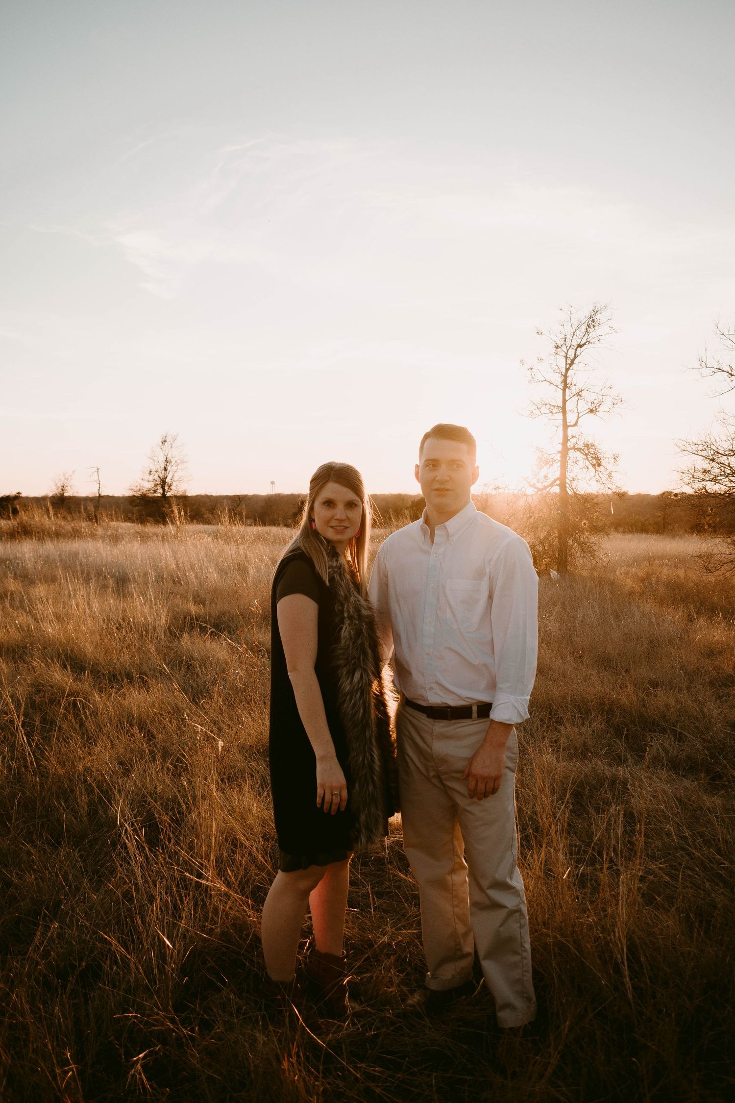 Couple poses together in casual attire during sunset in a rustic field setting.
