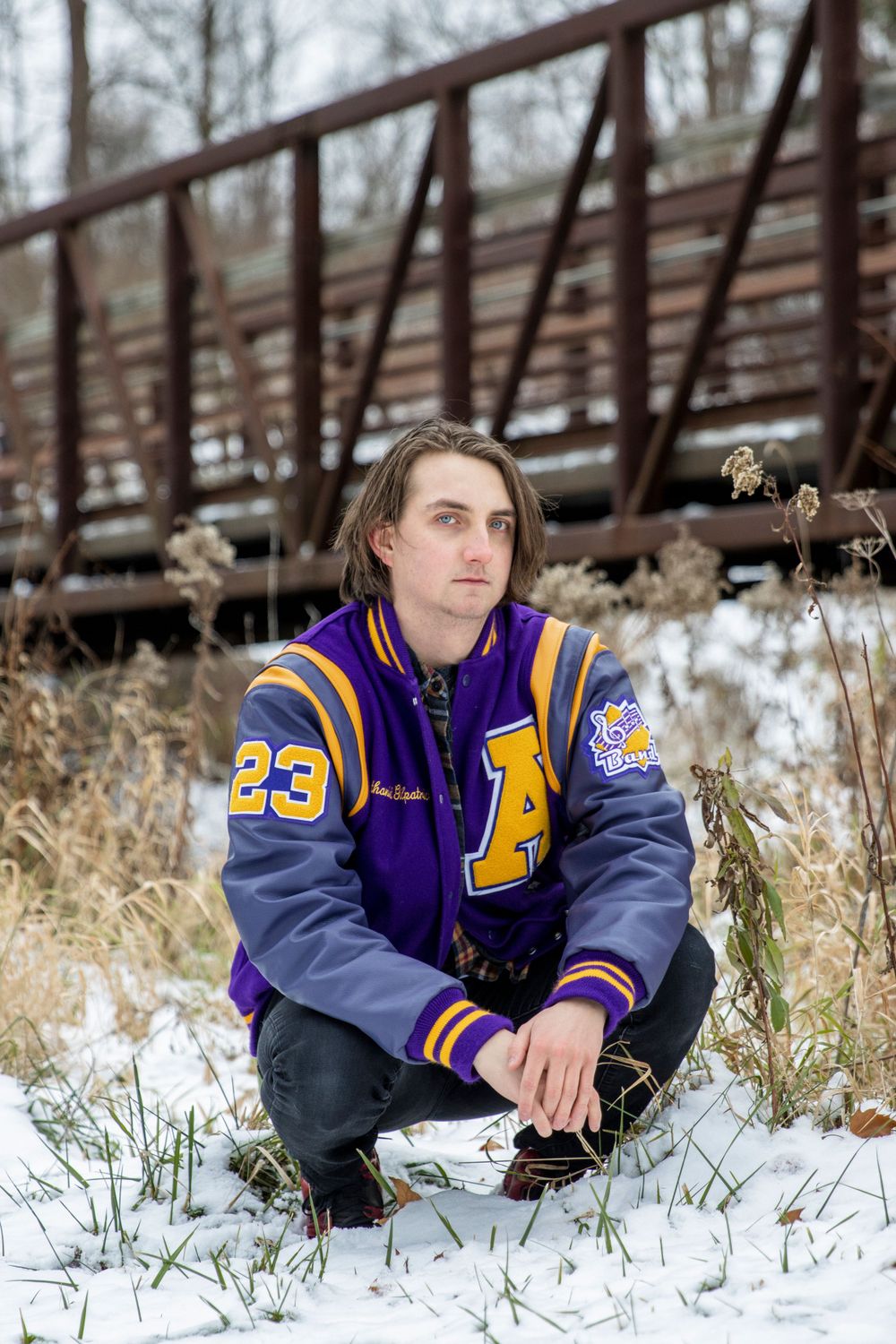 senior boy in his varsity jacket squatting in front of a red bridge on a snowy day in utica
