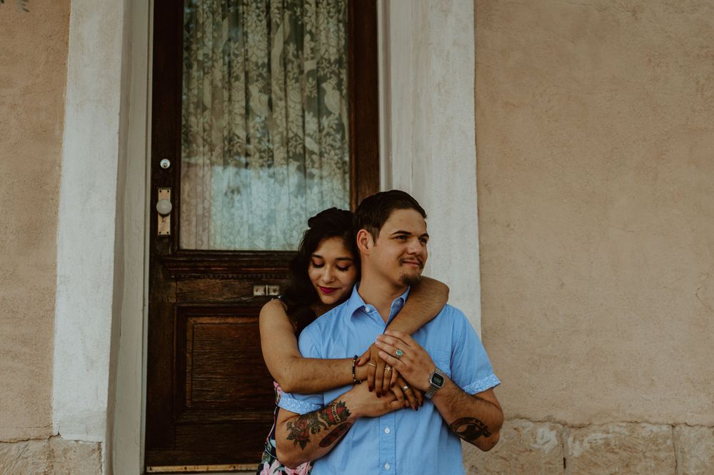 A couple shares an intimate moment on stone steps against a white stucco wall with wooden doors.
