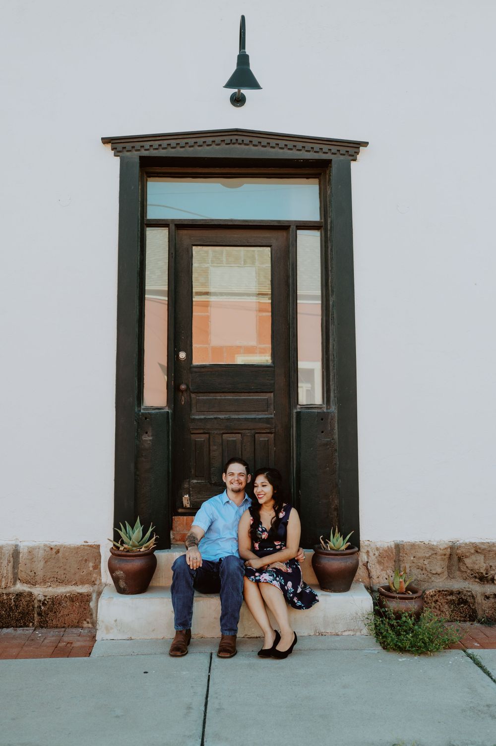 Two people sit together by dark wooden doors with clay planters on concrete porch.