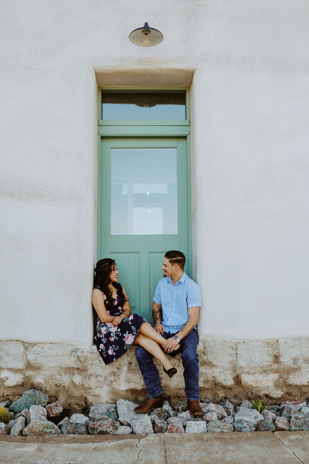 Couple sitting on stone ledge against white wall with turquoise door having intimate conversation.