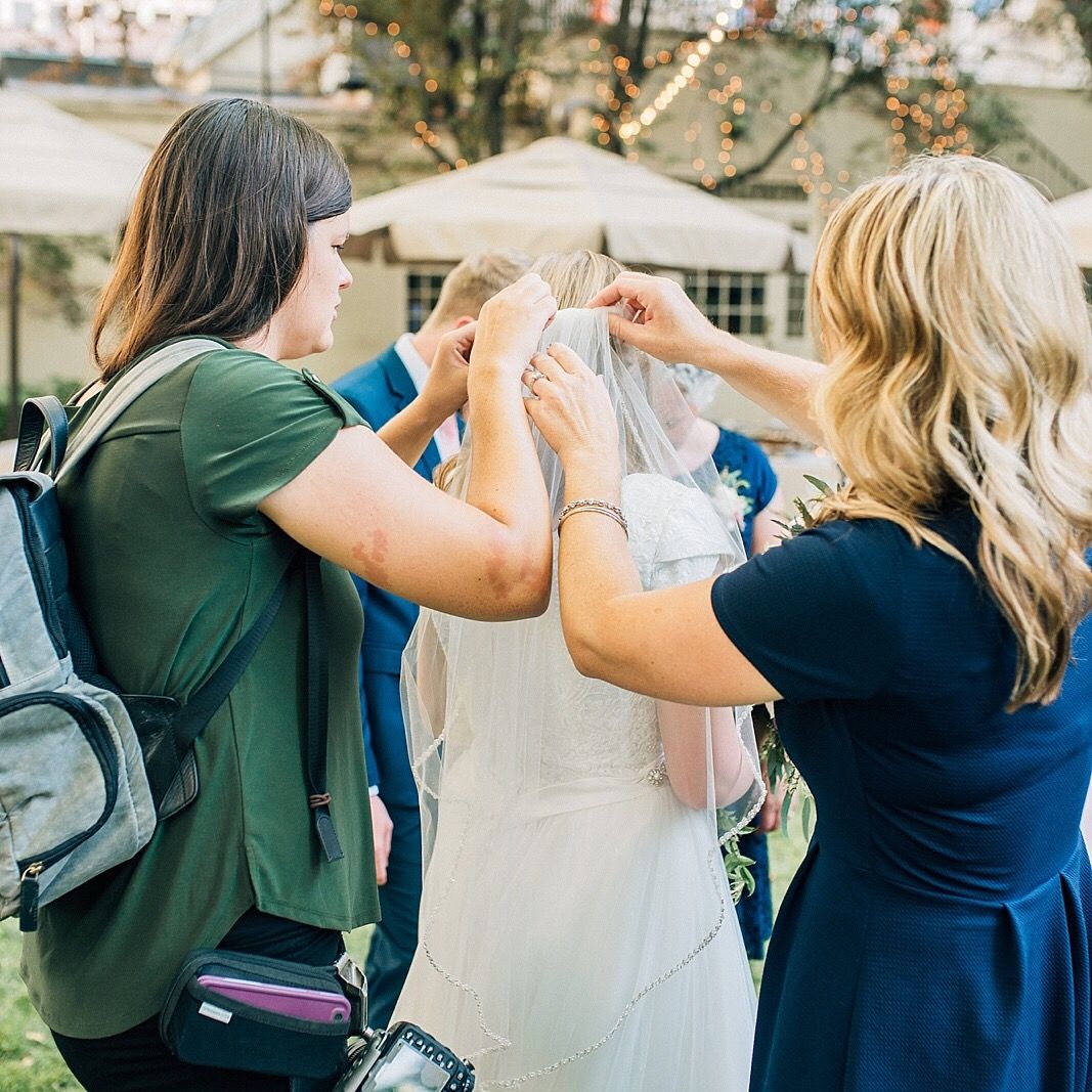 Two people help adjust a white wedding veil during wedding preparations outdoors.