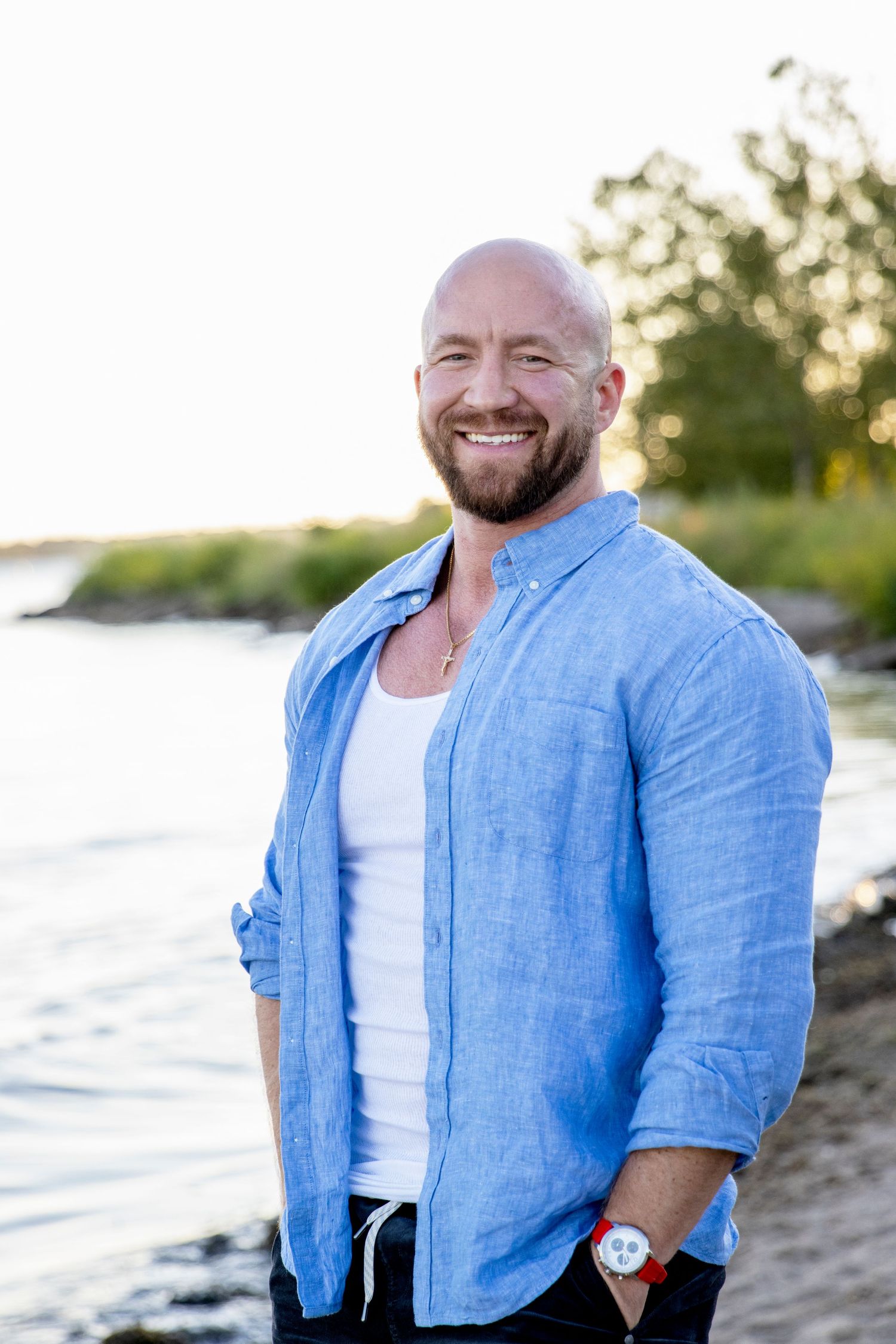 A man in a blue linen shirt and white tank top smiles warmly while standing on a beach of lake st Clair at sunset.