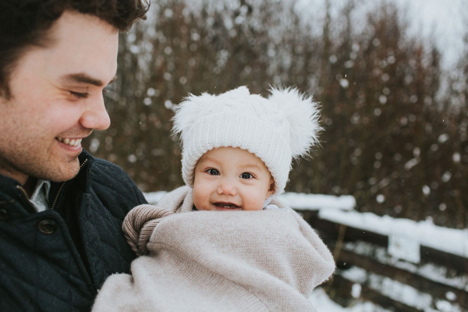 A smiling toddler wrapped in a blanket and is held by her dad in the snow.