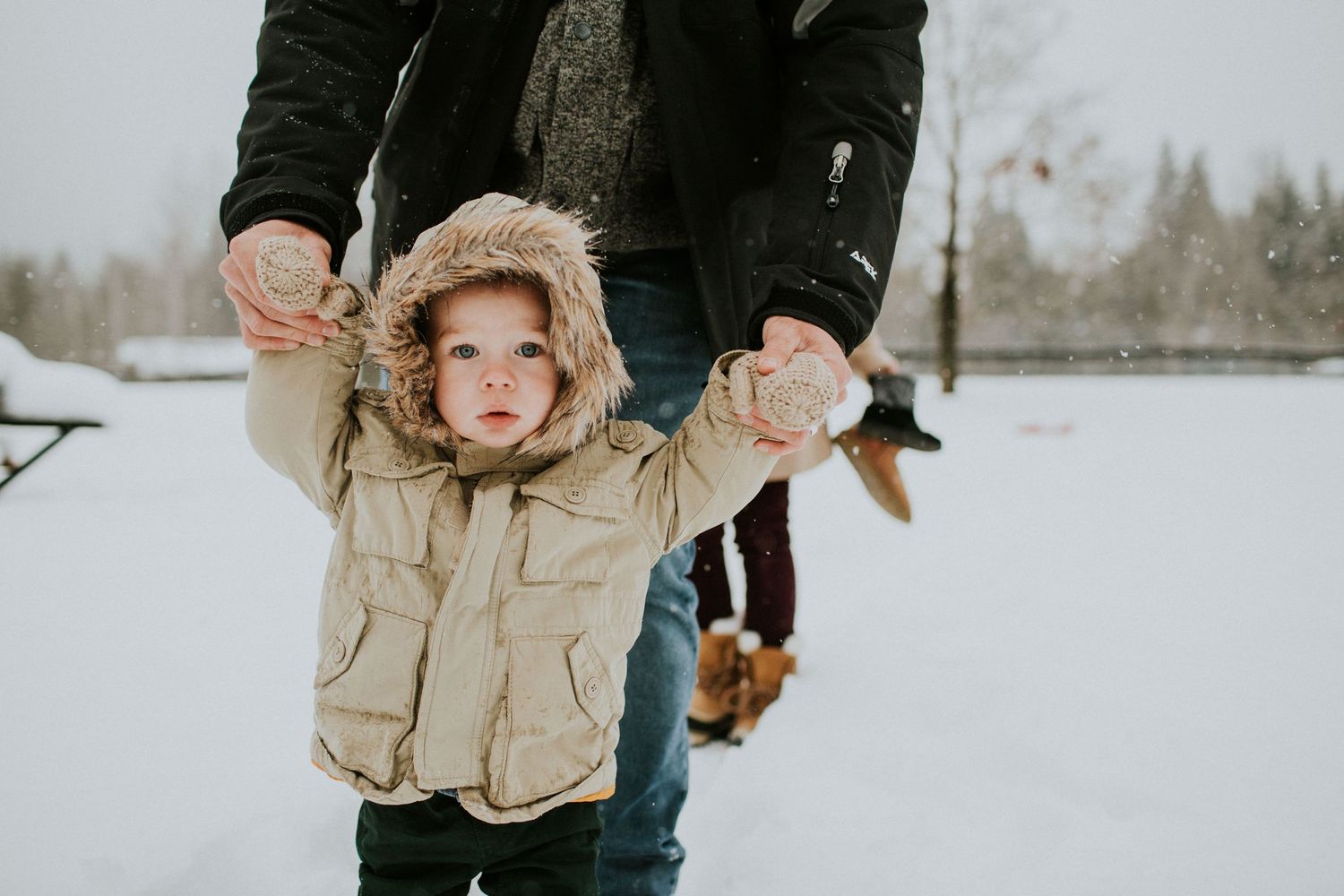 Small child wearing a fur-trimmed hooded winter coat holds adult's hands while standing in snowy landscape.