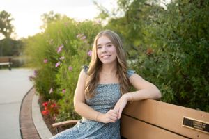 Young person in floral dress sits on wooden bench in garden setting at sunset.
