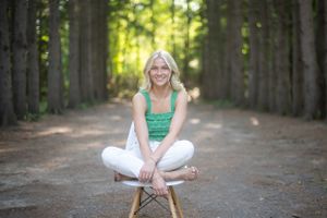 A person sits cross-legged on a forest path lined with tall evergreen trees.