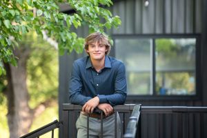 Teen leans on black railing in casual setting with natural backdrop.