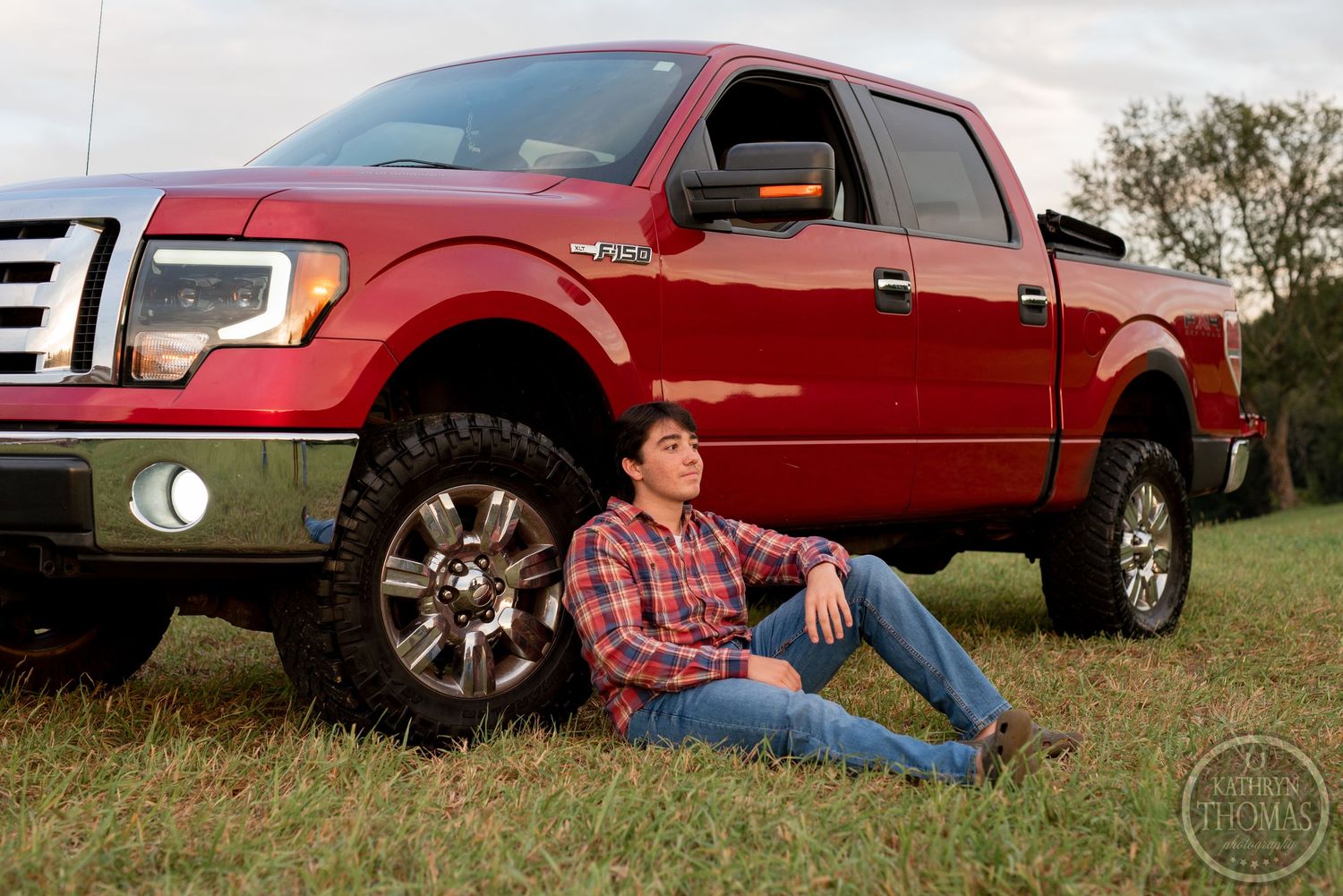 Person in plaid shirt poses beside red Ford F150 pickup truck in field.