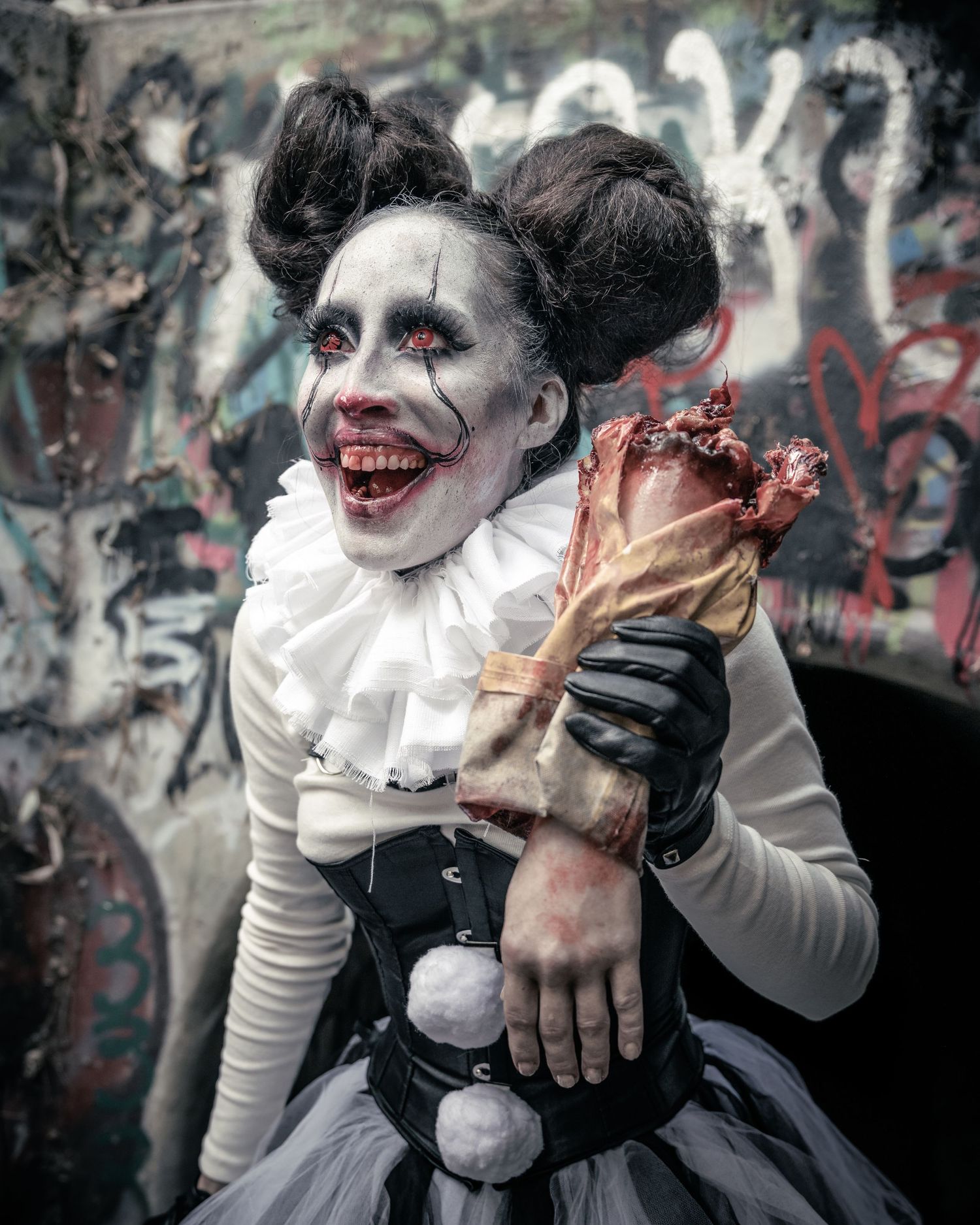 Female model as Pennywise the clown in a graffiti-covered sewer, holding a severed hand prop with red eyes and subtle blood effects.