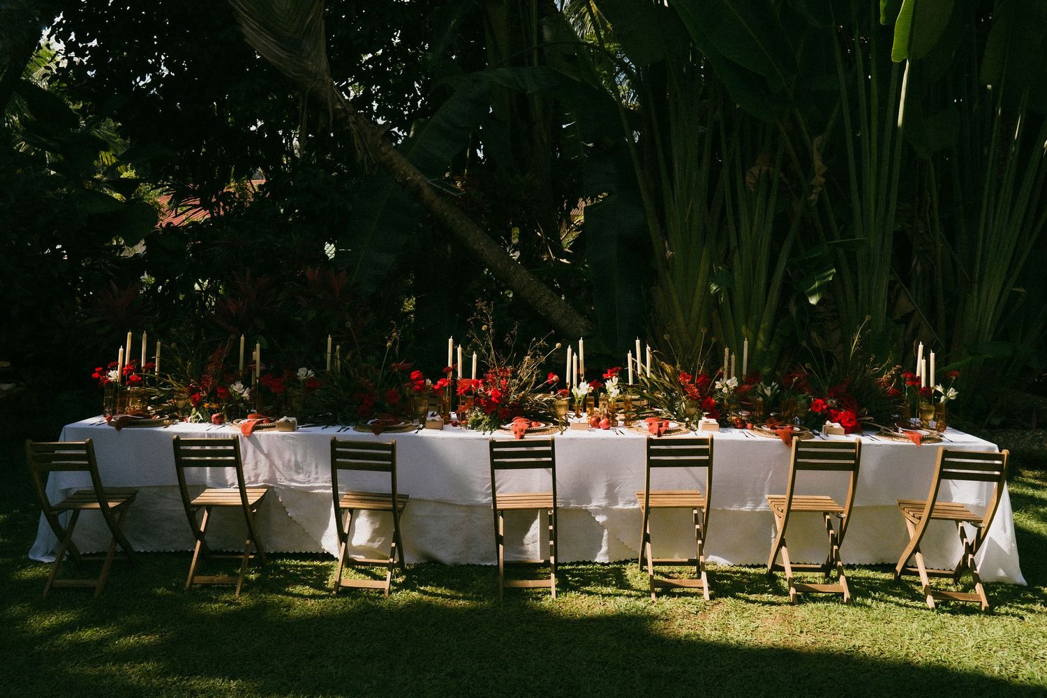 Elegant outdoor wedding reception table with red floral centerpieces and candles set in a lush tropical garden.