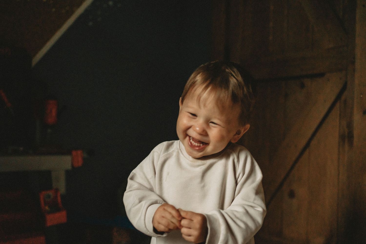 A toddler in a white sweater laughs joyfully in a rustic indoor setting.