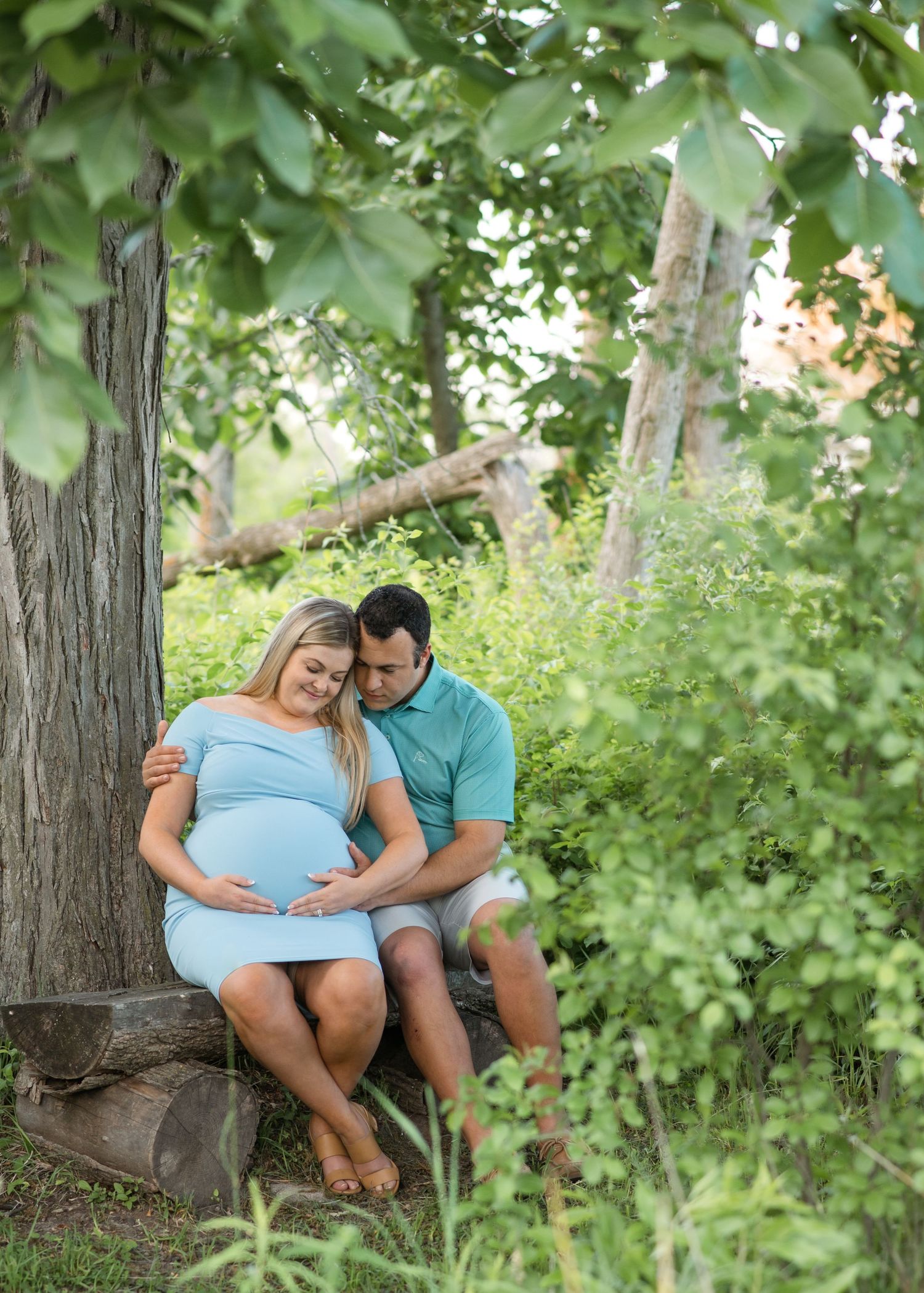 Expectant couple shares intimate moment while sitting on tree trunk in lush green setting.