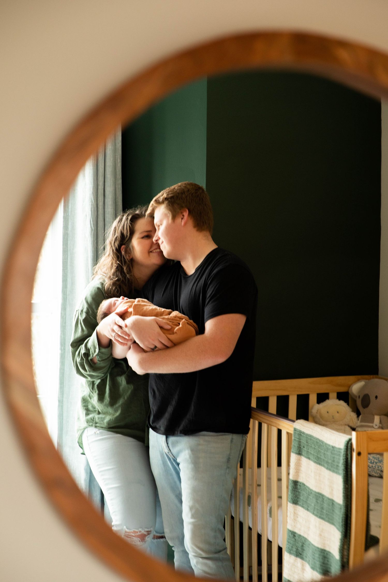 Tender moment between parents holding newborn baby by window in nursery with round mirror frame.