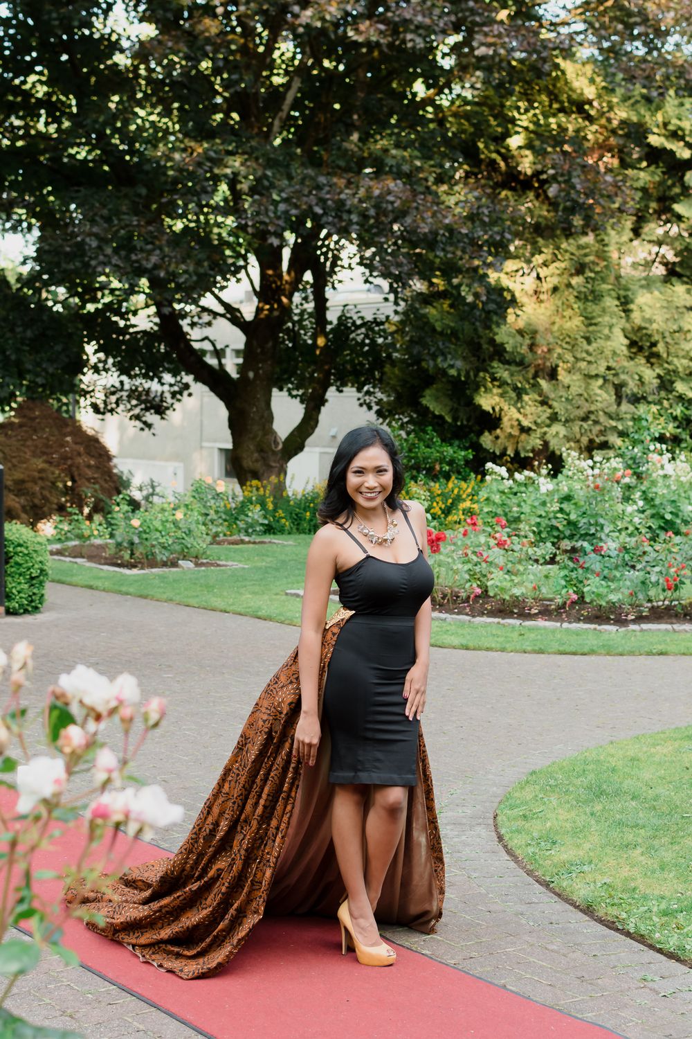 A woman poses on a red carpet outside amongst the trees in a rose garden.