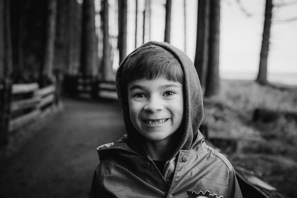A black and white image of a young boy smiling cutely at the camera while he walks outside along a forest path.