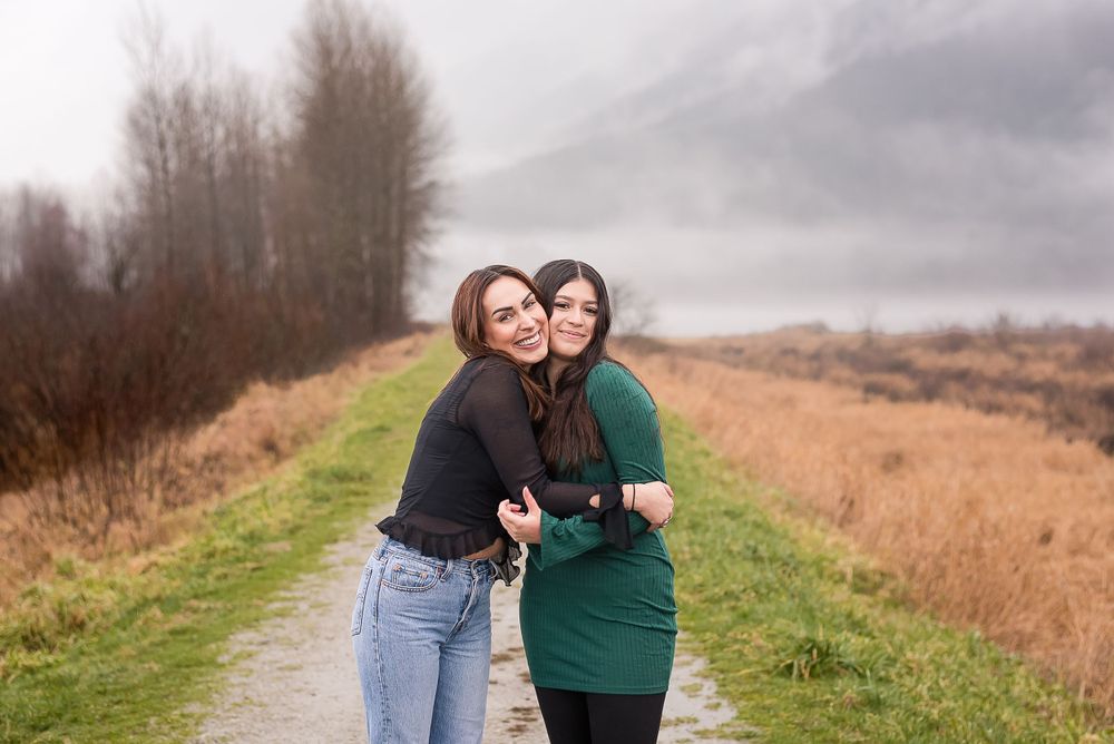 Two sisters embrace one another in the rainy winter outdoor landscape. Mountains and fog feature in the background of Pitt Lake, BC.