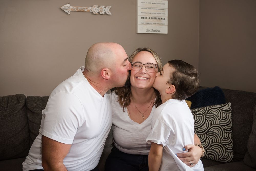 A father and son kiss mom on the cheek during an in-home family photo session, while she giggles away.