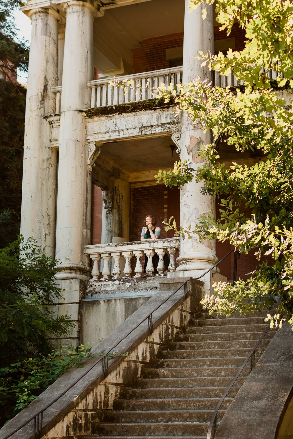 At a historic abandoned building, a woman looks out from the balcony to the grand staircase and green bushes below.