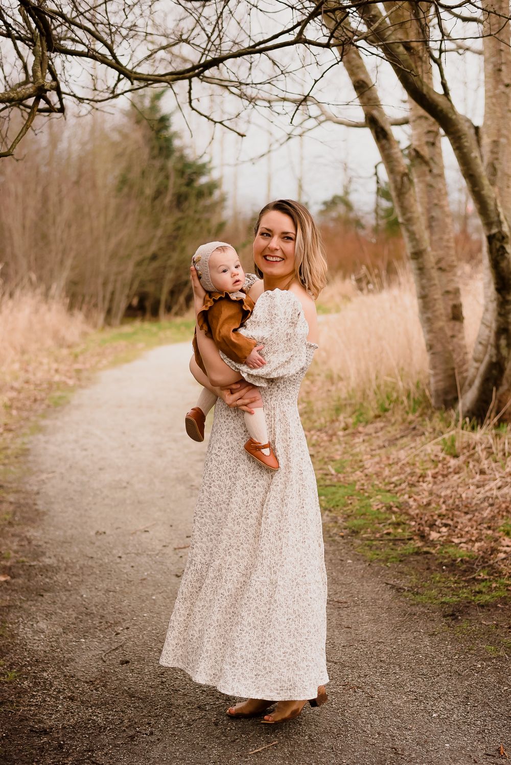 On a tree lined path a mother dressed in a neutral dress holds her baby daughter wearing a bonnet.
