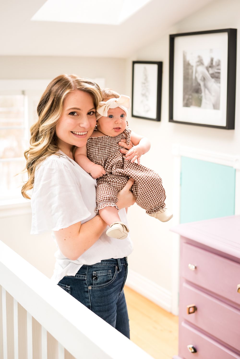A proud mother stands inside a beautifully lit home holding her baby daughter who is wearing a bow on her head.
