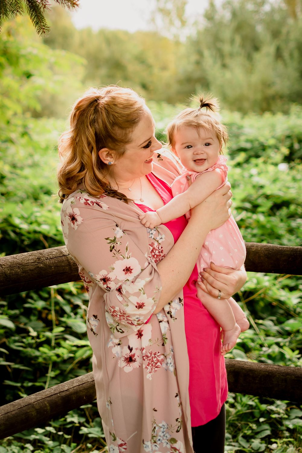 A mother and her baby daughter in beautiful pink outfits stand in the spring sunshine, smiling.