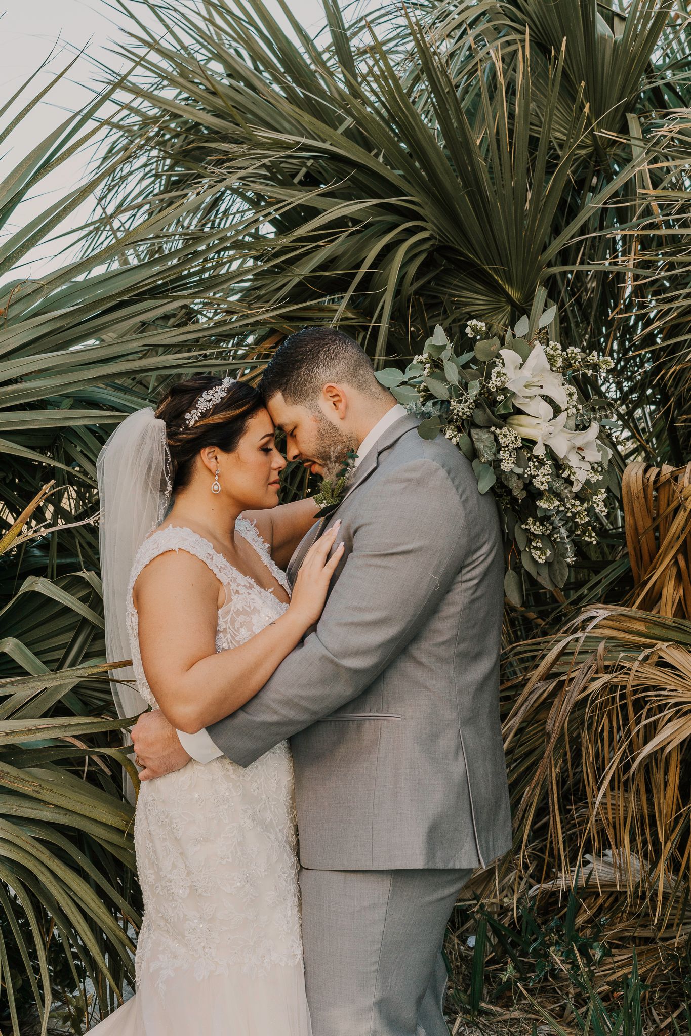 Intimate wedding portrait amidst tropical plants with bride in white lace and groom in grey suit.