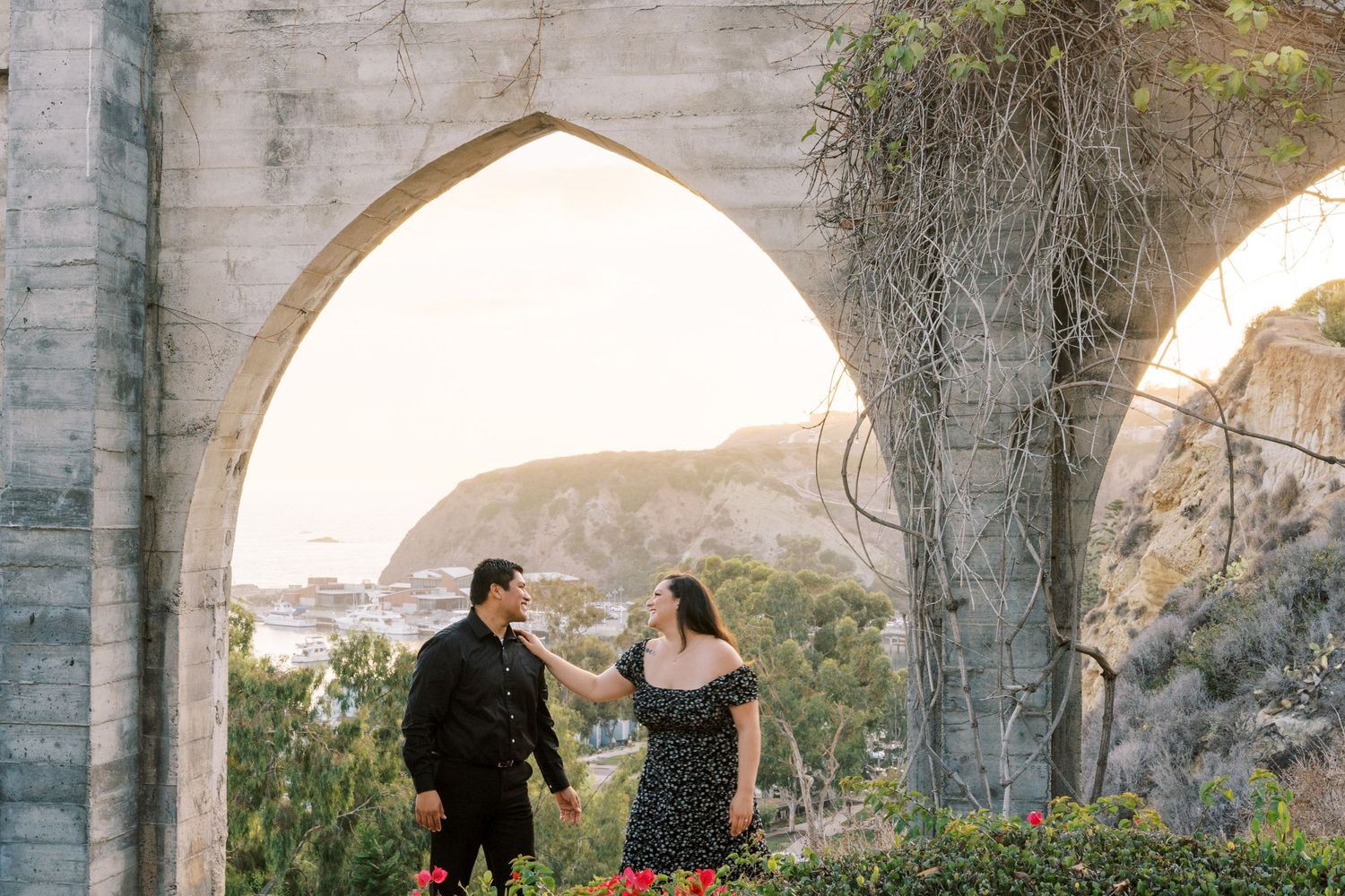 Couple sharing romantic moment under stone archway with mountain vista behind them.
