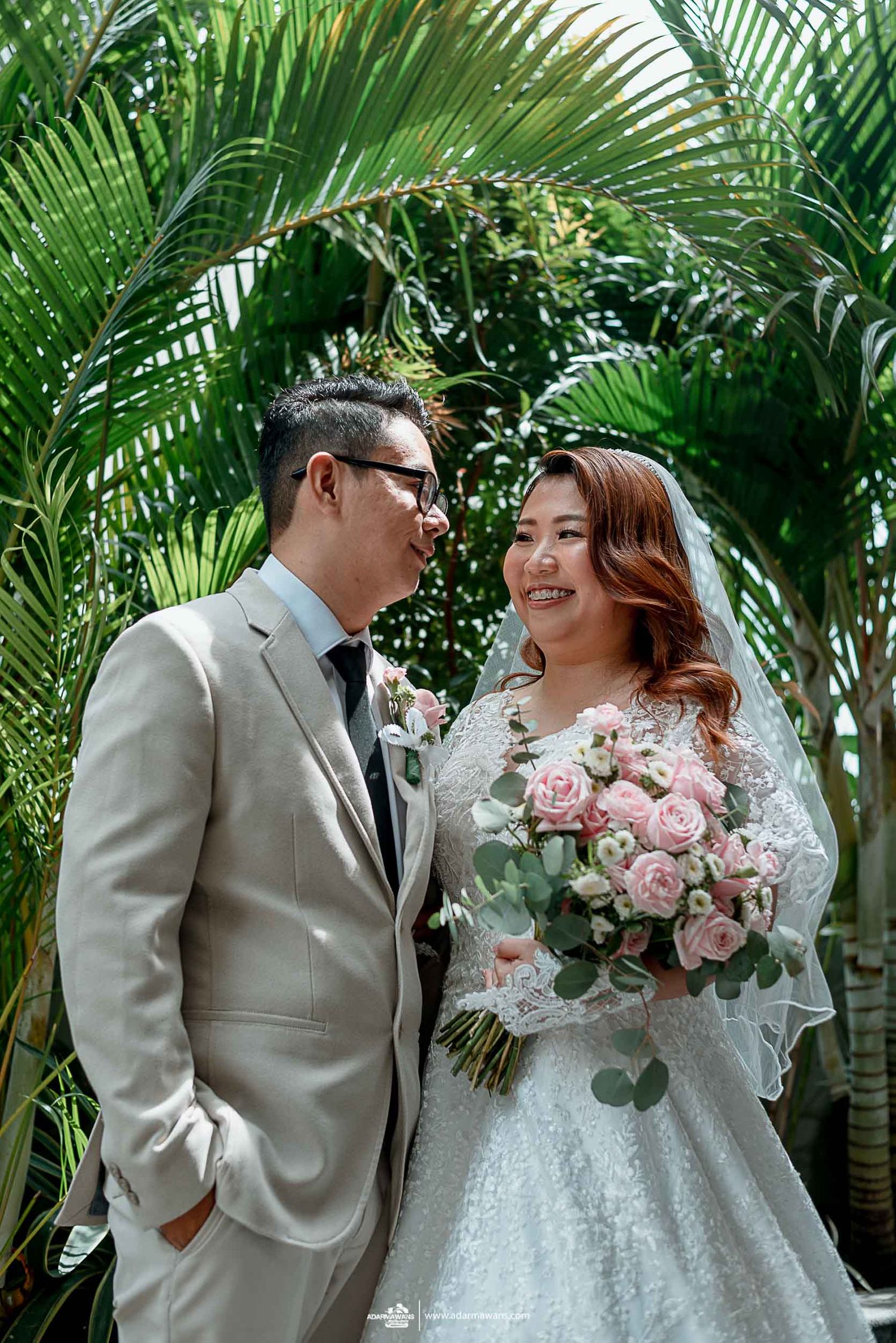 Couple in wedding attire share intimate moment among lush palm garden setting.