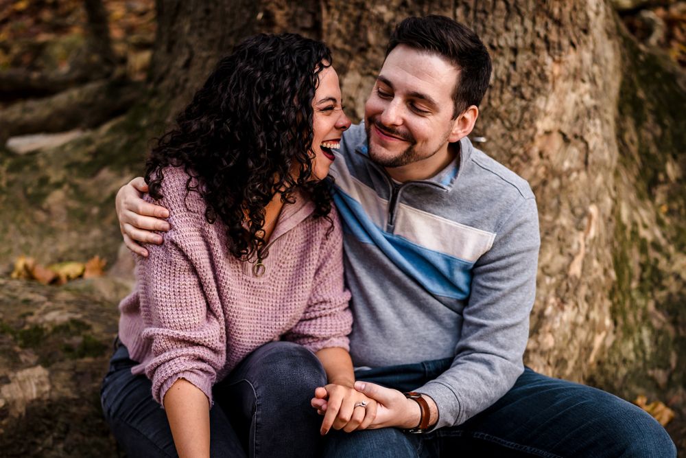 Couple sitting together against a large tree trunk surrounded by fallen autumn leaves.