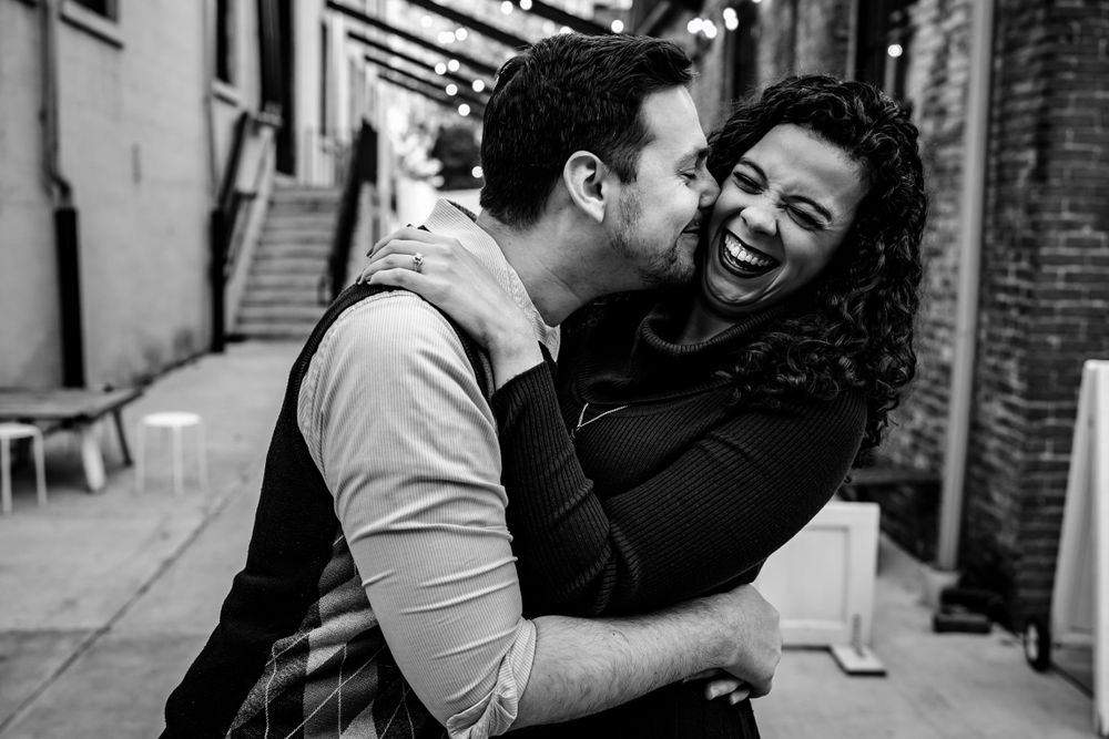 A couple shares a joyful embrace and laugh together in a black and white urban setting.