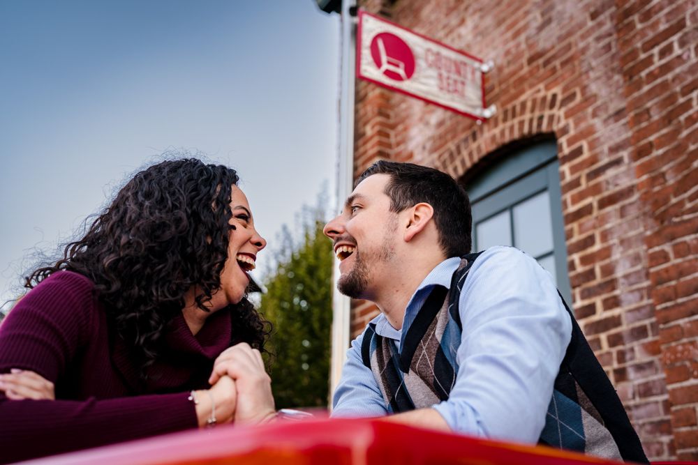 A couple shares a candid laughing moment against a red brick wall background.
