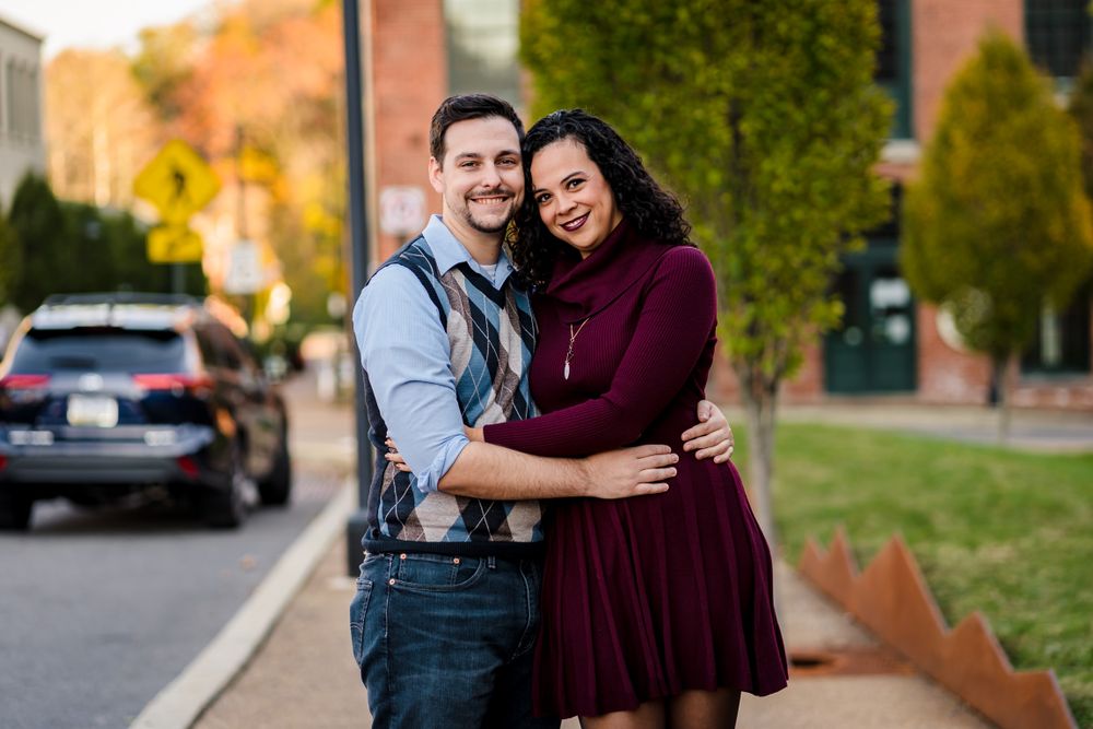 A couple poses together outdoors in autumn with brick buildings and fall foliage in the background.