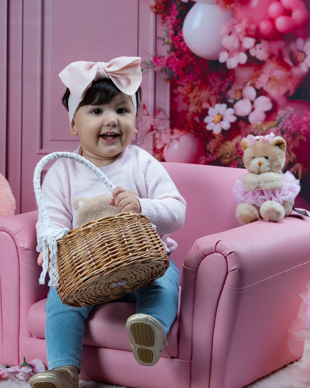 Smiling baby holding wicker basket on pink armchair with floral backdrop.