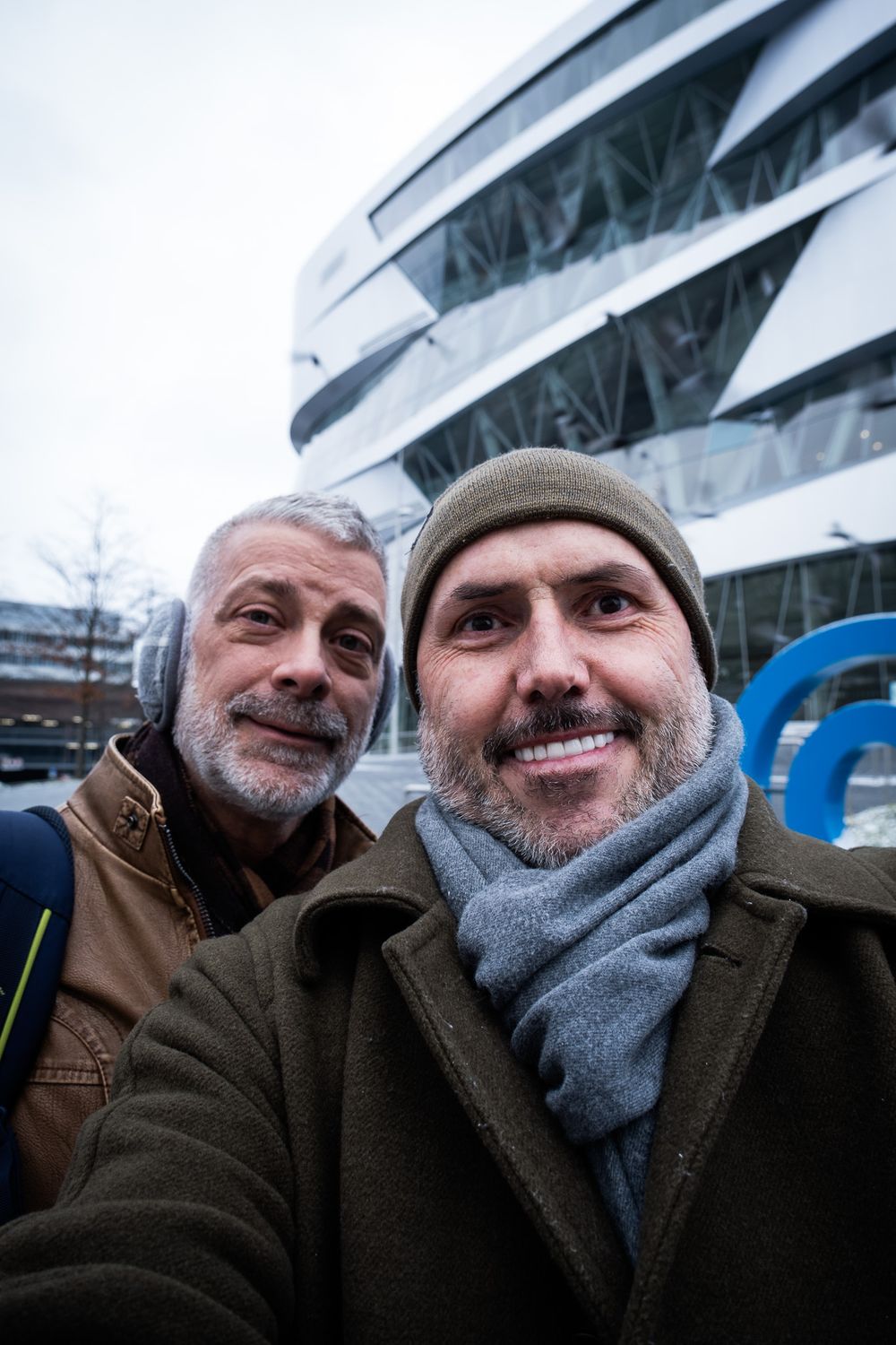 Two smiling men taking a selfie outside the Mercedes-Benz Museum in Stuttgart on a winter day.