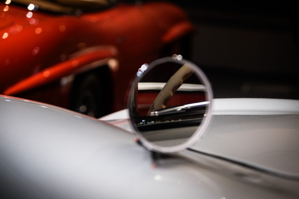 Reflection of a red vintage car captured in the round chrome side mirror of a classic white automobile.
