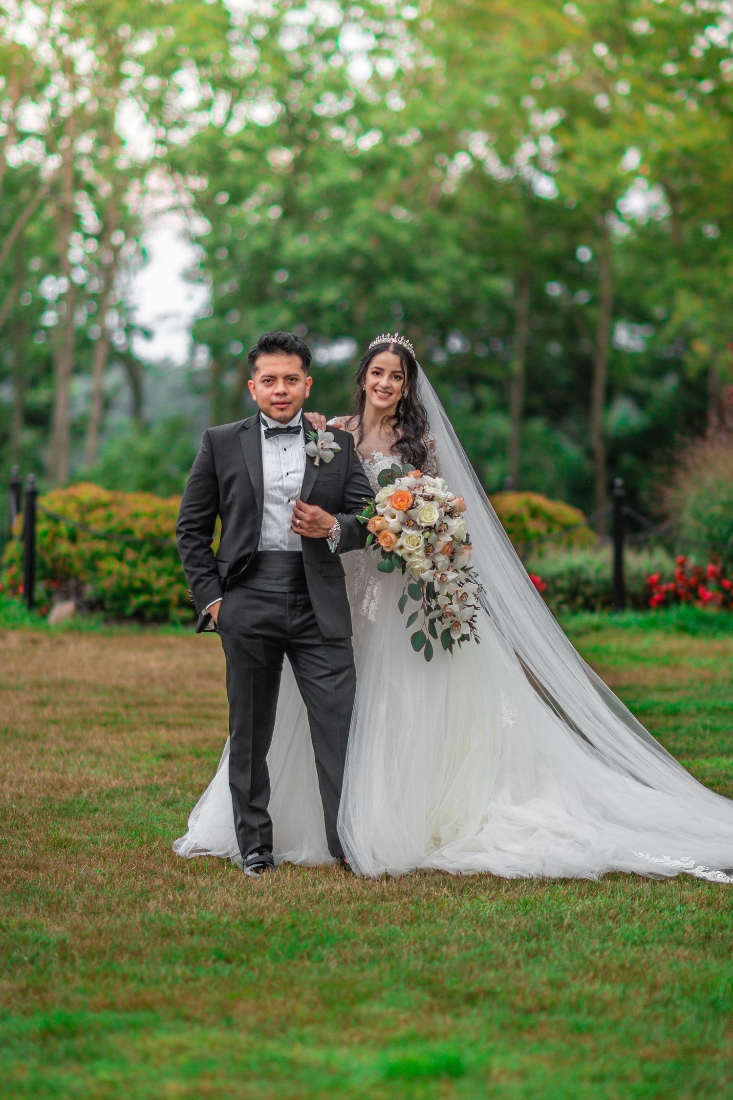 A wedding couple poses in a lush garden setting with elegant attire and flowing veil.
