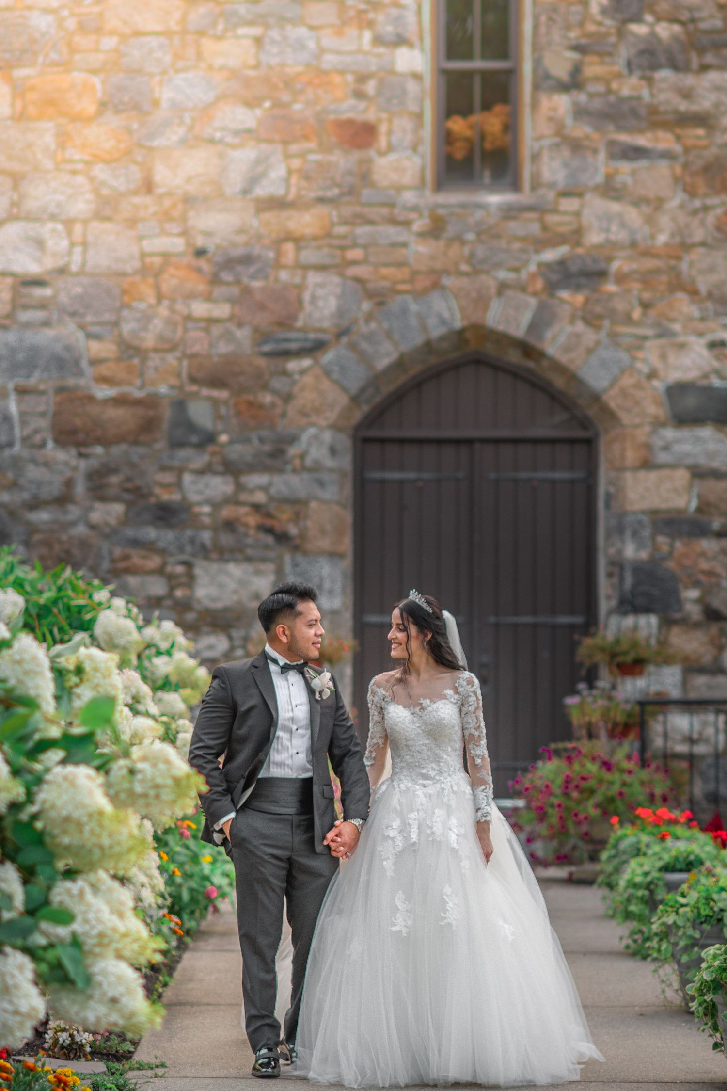 A couple walks hand in hand along a stone church path surrounded by hydrangeas.