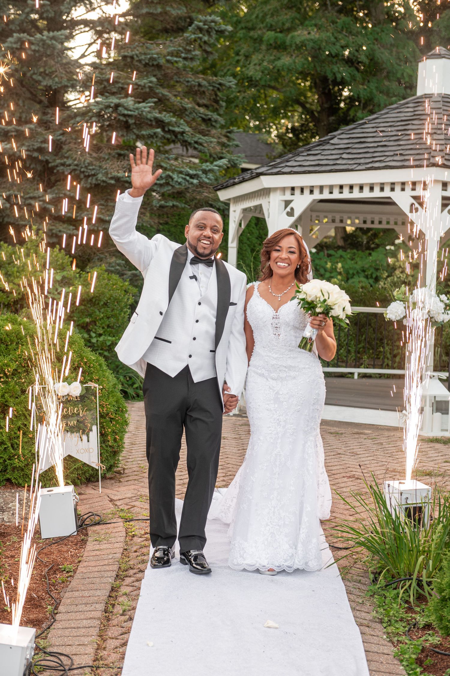 Wedding couple walking along brick pathway by white gazebos in garden setting.