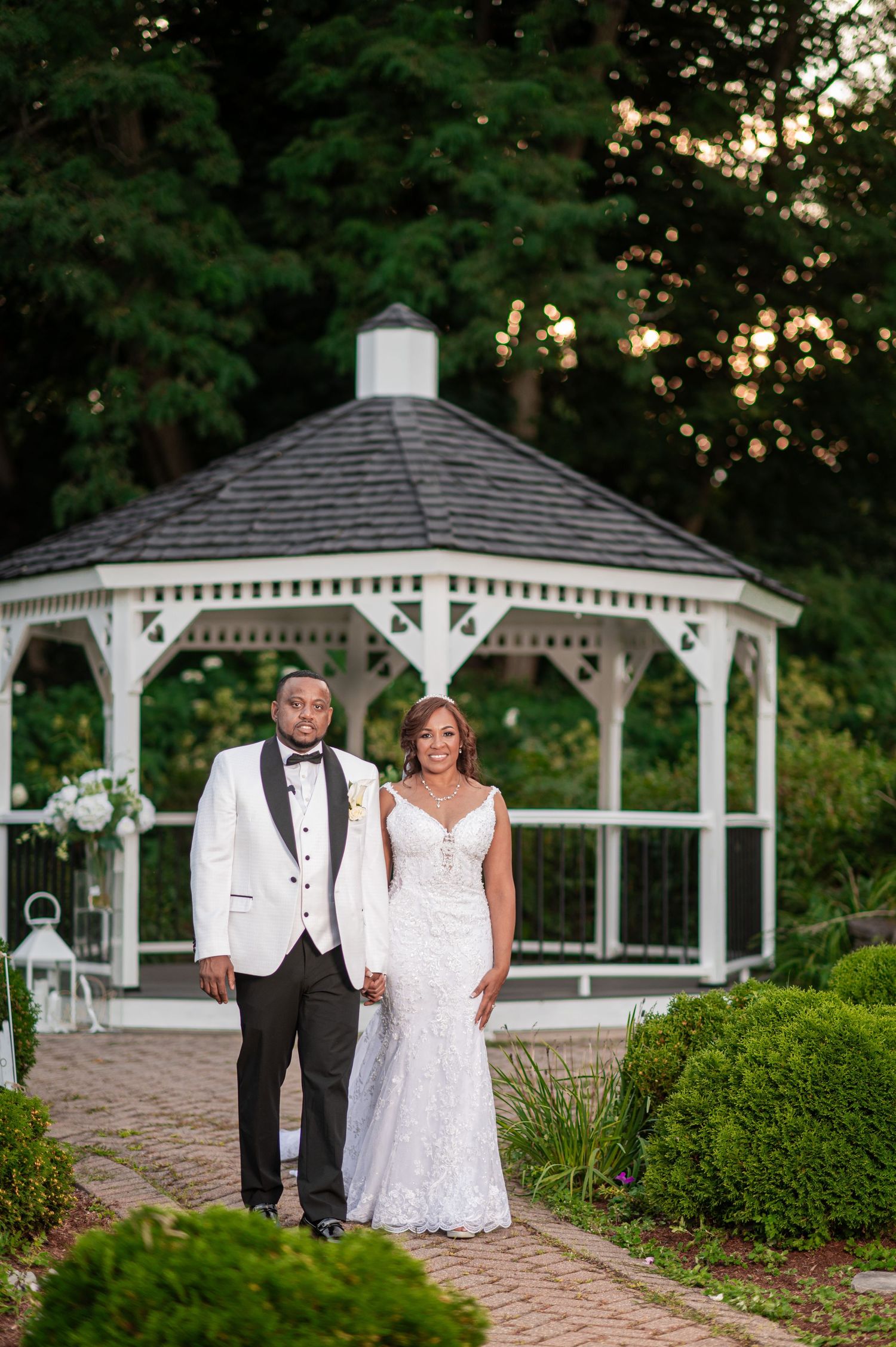 Elegant wedding photos near white gazebos on brick pathway with manicured gardens.