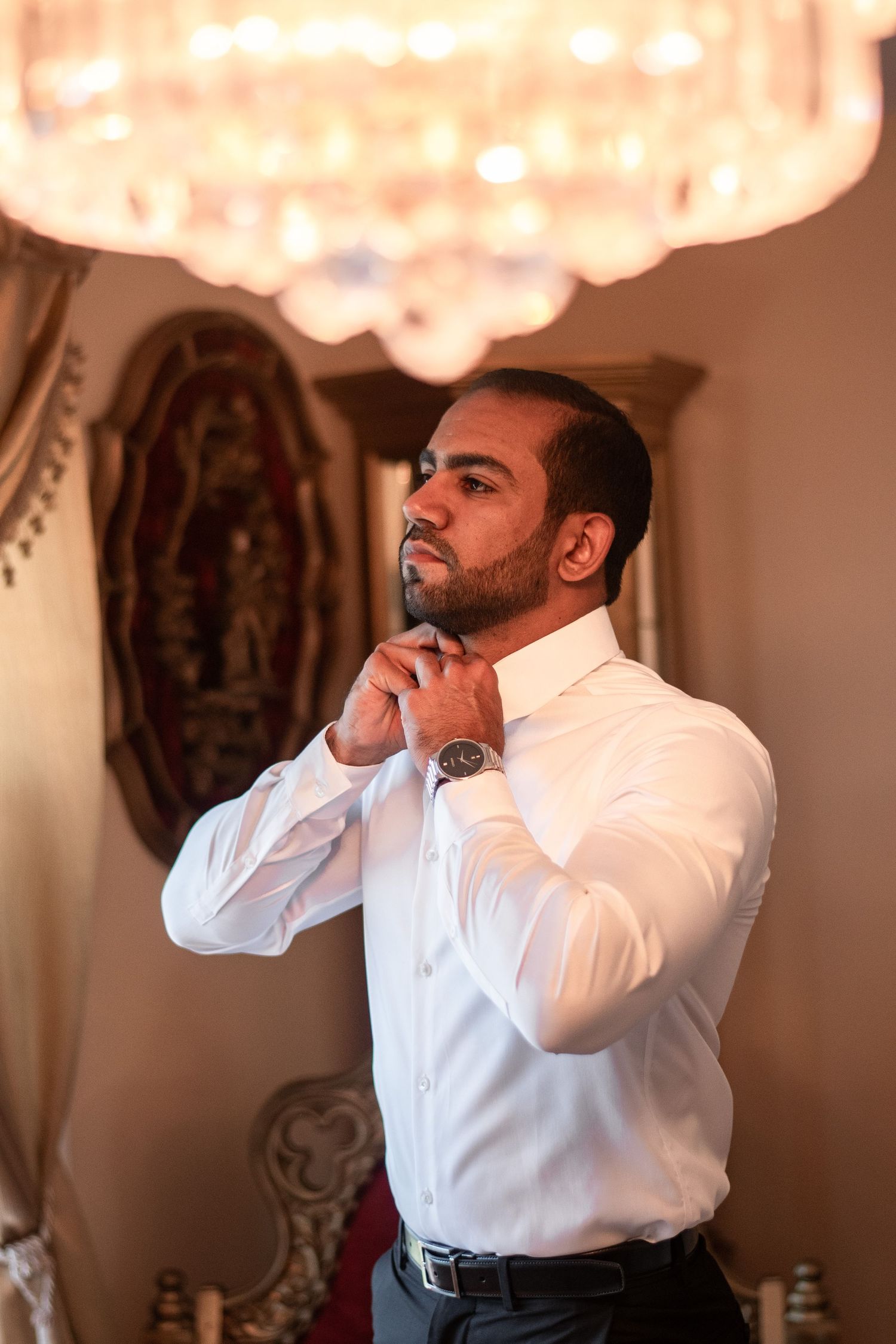 Formal portrait of person adjusting white shirt collar under crystal chandelier.